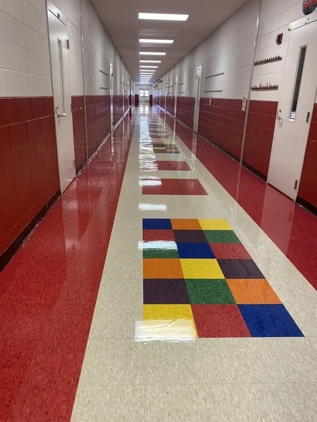 A school corridor with red vinyl down both sides, a cream centre lane and a patchwork of blue, orange, green and gold tile squares, all burnished to a gloss