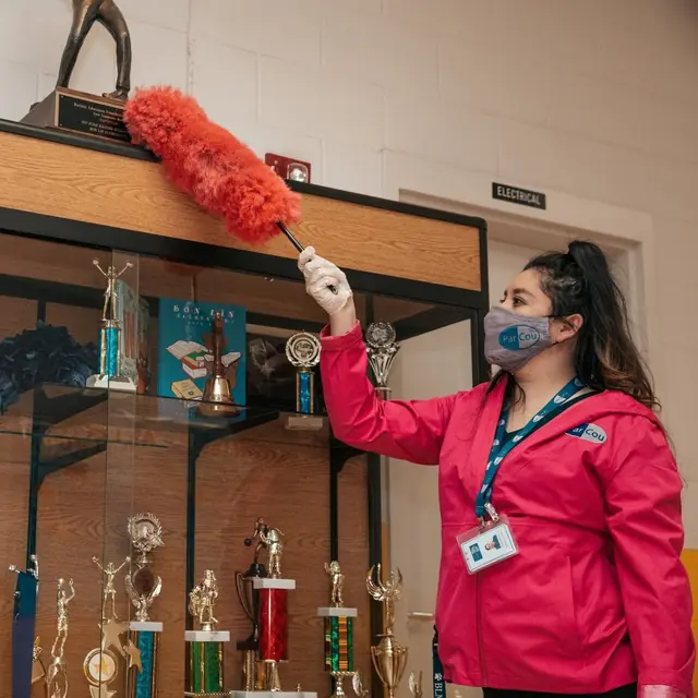 A ParCou team member in a face mask and lanyard dusting the top of a school trophy case with a red duster